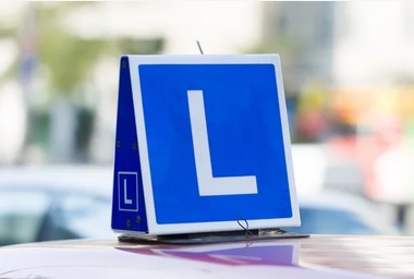 Niebieska tabliczka z dużą białą literą L umieszczona na dachu samochodu, sygnalizująca naukę jazdy, z rozmytym tłem miejskiego ruchu drogowego